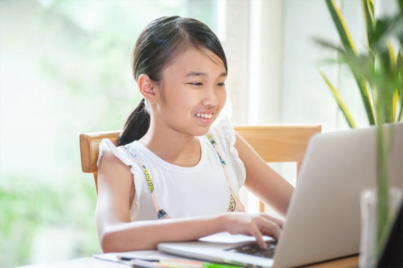 a young girl seated behind a laptop and smiling while participating in homeschooling